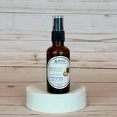 samhain room and linen spray Brown spray bottle labeled 'Brigid' on a white pedestal against a wooden background