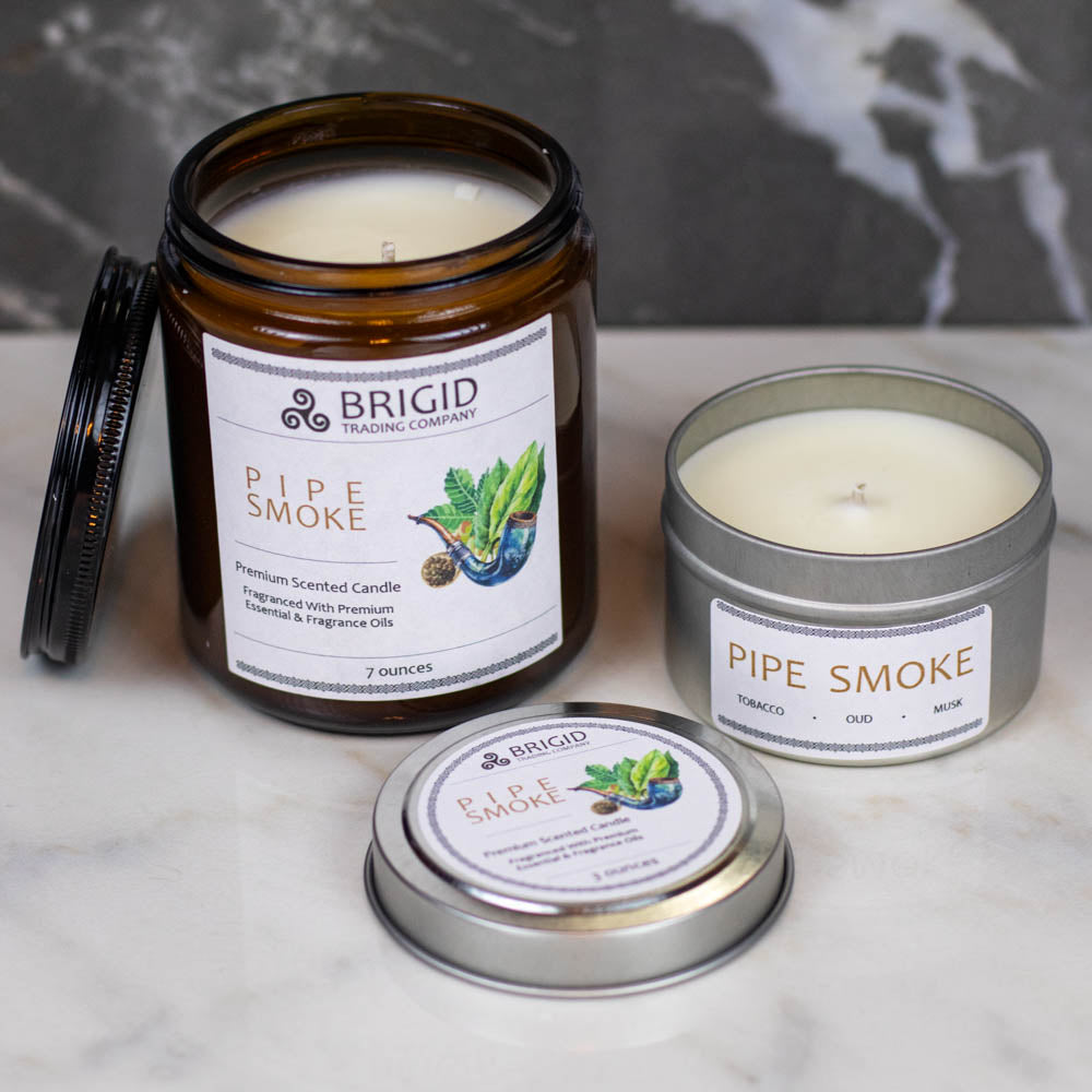 pipe smoke premium soy wax candle by brigid trading company tobacco smoke oud wood and musk photo shows a dark marble background with light marble foreground and two candles one amber glass jar candle one tin small container candle 