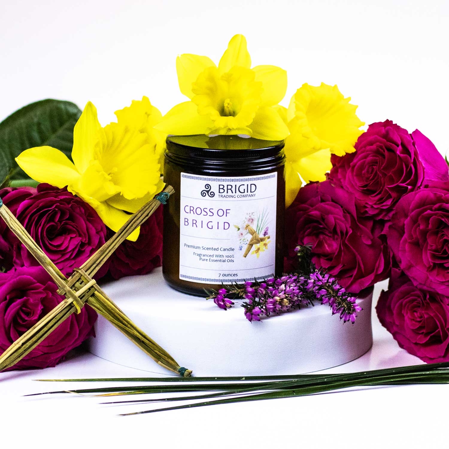 cross of brigid candle with flowers lifestyle shot image is white with candle in the center and a st brigids cross made of reeds, roses, daffodils narcissus, vetiver, and pink heather fresh flowers by brigid trading company llc