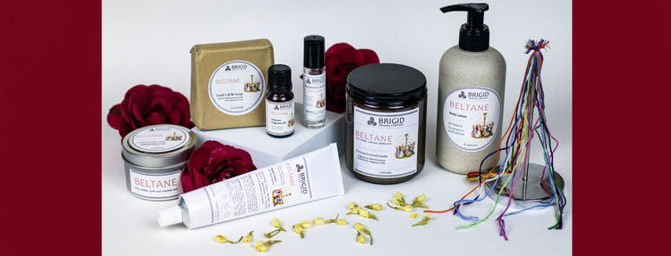 beltane 2025 collection image for brigid trading company llc kitsap county washington state spring summer collection rose jasmine scented celtic holiday fragrance collection image shows white background with beltane soy wax candle, goats milk soap, shea butter hand lotion, fragrance oil, diffuser oil, perfume oil, roller bottle perfume, rose, shea butter natural body lotion, beltane maypole with jasmine flowers and red roses
