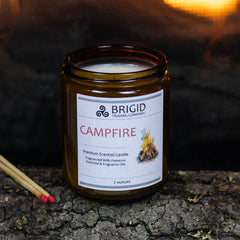 campfire mesquite smoke warm woods scented candle for camping fans mens gifts womens gifts for her for him outdoor fan presents hiking is in tents hand made in kitsap county independent american owned business usa grown sustainable soy wax not tested on animals original studio image by brigid trading company photography image shows one large candle seven ounce jar candle in amber glass with fire in background, matches and wooden log