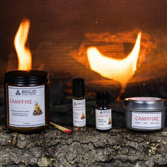 campfire mesquite smoke warm woods scented candle for camping fans mens gifts womens gifts for her for him outdoor fan presents hiking is in tents hand made in kitsap county independent american owned business usa grown sustainable soy wax not tested on animals original studio image by brigid trading company photography image shows two candles large and small with roller bottle and diffuser oil bottle full collection gift set fire in background, matches and wooden log