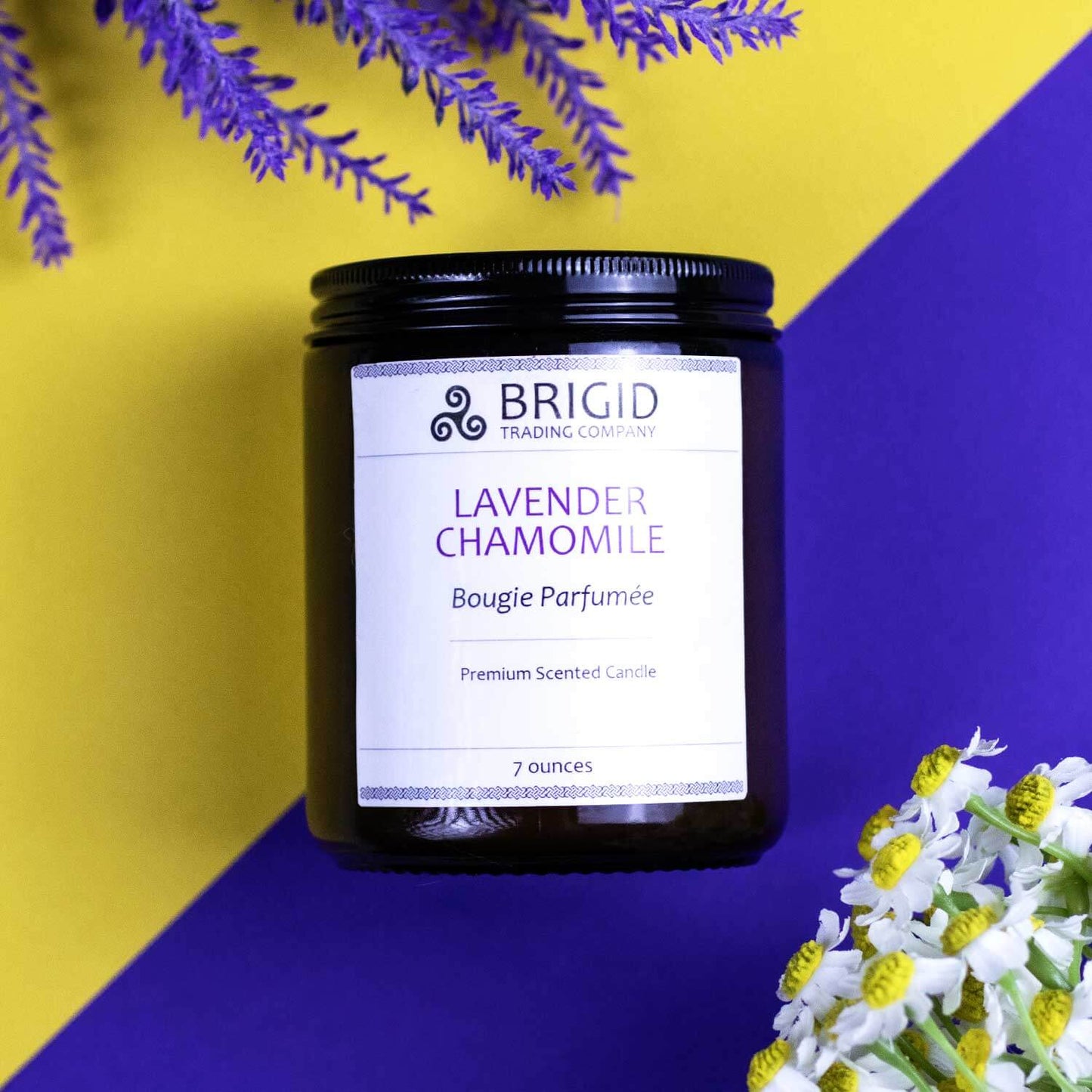 lavender chamomile bougie parfumee premium scented candle by brigid trading company soy wax hand poured hand made glass jar candle background is bright vivid purple and yellow product is in the center with contrasting flowers on opposite corners