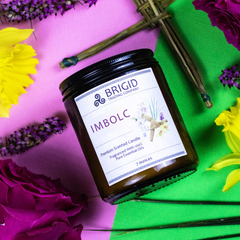 imbolc holiday candle cross of brigid by brigid trading company imbolc candle rose narcissus daffodil st brigids cross reeds vetiver heather spring collection celtic irish holidays midwinter february witchy modern christian pink and green background vivid cross and ingredients as props surrounding candle original photography studio shot by brigid trading company llc