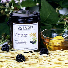 lughnasadh candle 7oz by brigid trading company llc blackberry oat honey scent premium soy wax candle photography original by brigid trading company blackberry leaves and plants in background with fresh blackberries sitting on dried oats with bowl of honey and honey spoon next to candle in amber glass jar with lughnasadh on the label 2024 kitsap county washington state united states